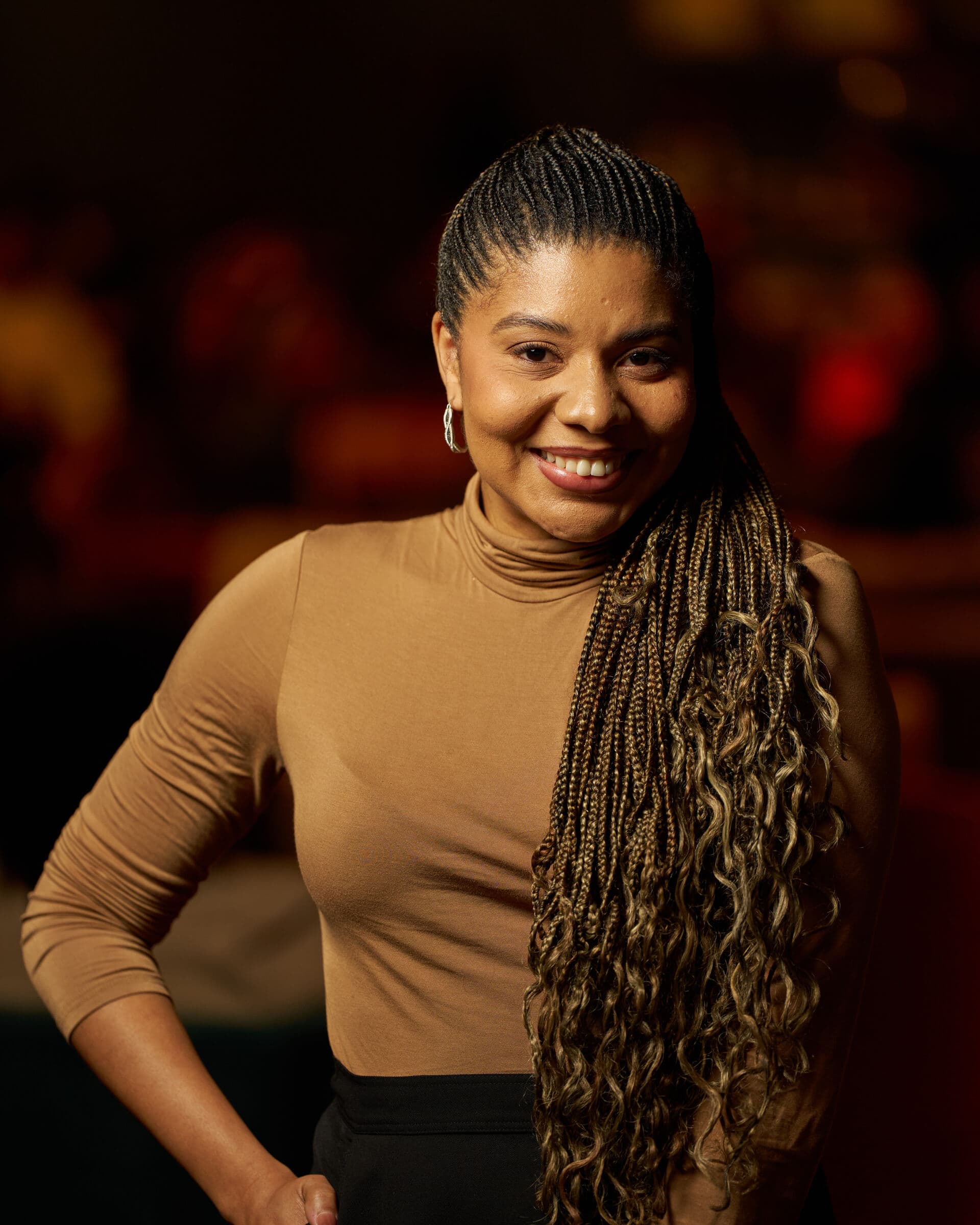 Jacqueline V. Twillie in a camel turtleneck and black skirt, one hand on her hip, against warm amber light.