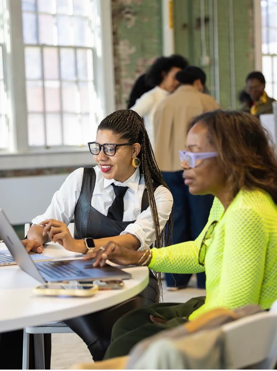 Jacqueline V. Twillie in a white shirt, black tie and leather vest, smiling as she works at a laptop beside an attendee at a CultureCon build session in Brooklyn.