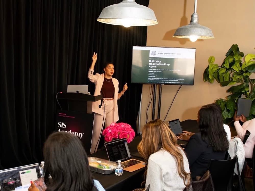 Jacqueline V. Twillie standing at the front of a Sistas In Sales Summit session, one arm raised, beside a screen that reads Build Your Negotiation Prep Agent, while attendees work at laptops.
