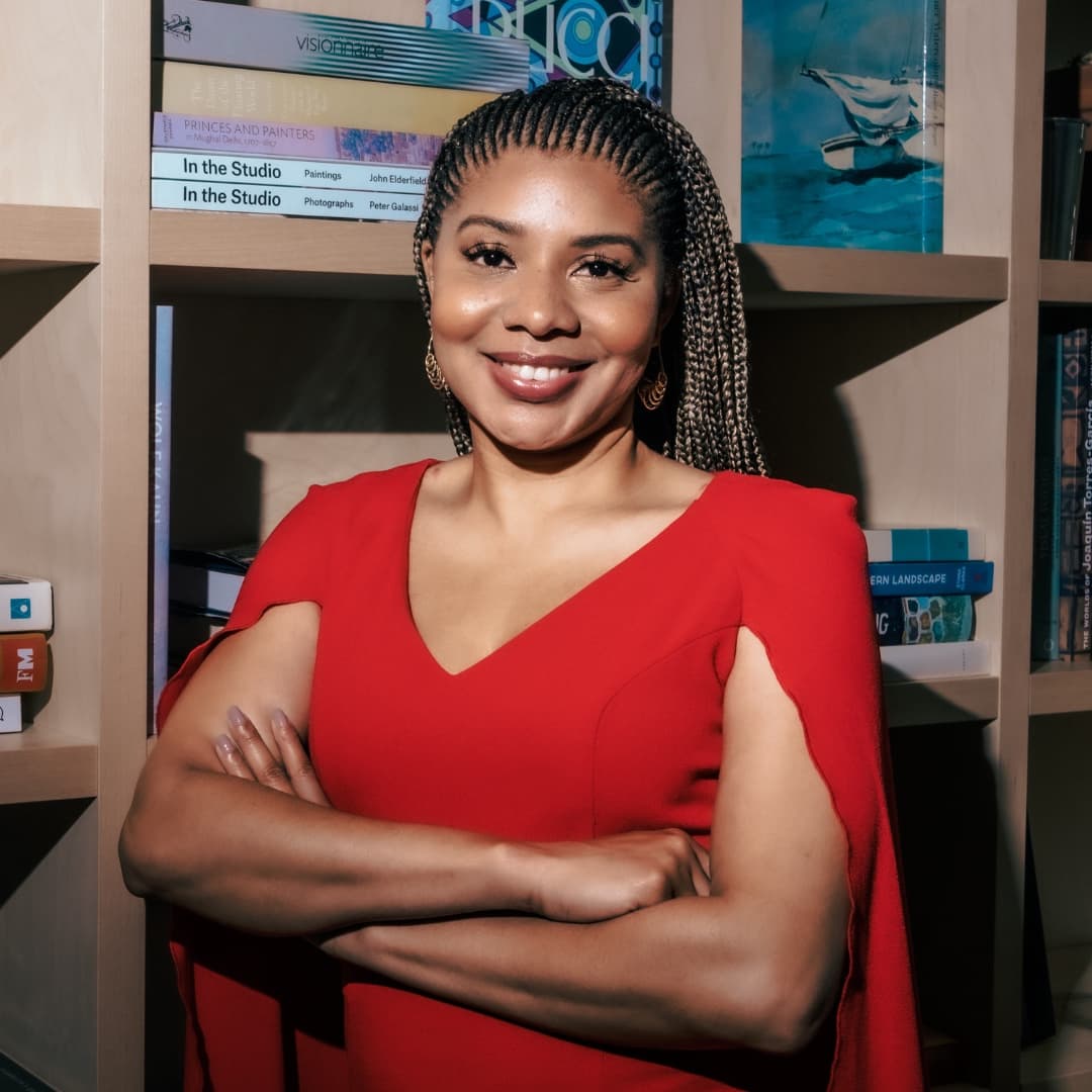 Jacqueline V. Twillie in a red cape dress, arms crossed, in front of a bookshelf.